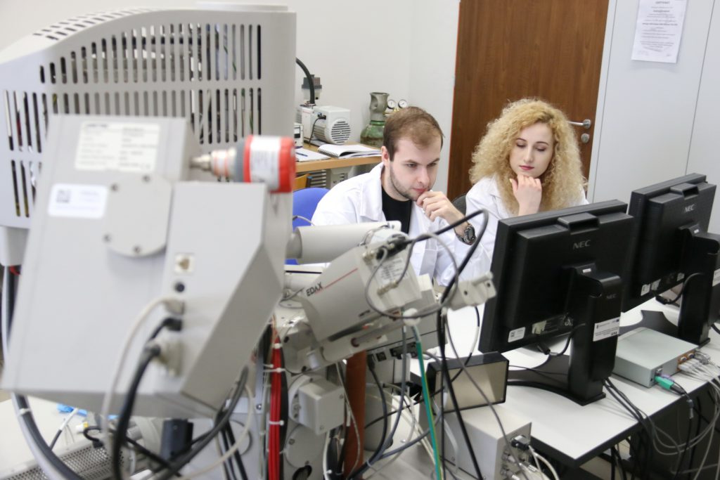 Dwoje naukowców w białych fartuchach pracuje przy skomplikowanej aparaturze laboratoryjnej i monitorach.
