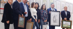 Grupa osób pozuje do zdjęcia z pracami plastycznymi na wystawie. Widać obrazy, rysunki i ramki.