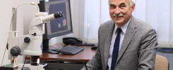 Mężczyzna w garniturze siedzi przy biurku z mikroskopem, monitorem i telefonem. Kontekst: laboratorium lub biuro.