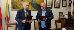 Dwóch mężczyzn w garniturach trzyma dokumenty. W tle obrazy, flagi i meble.
