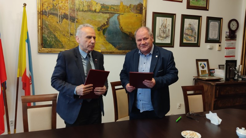Dwóch mężczyzn w garniturach trzyma dokumenty. W tle obrazy, flagi i meble.