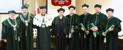 Ośmiu naukowców w togach akademickich stoi przed godłem Polski. Kluczowe obiekty: naukowcy, togi, godło.