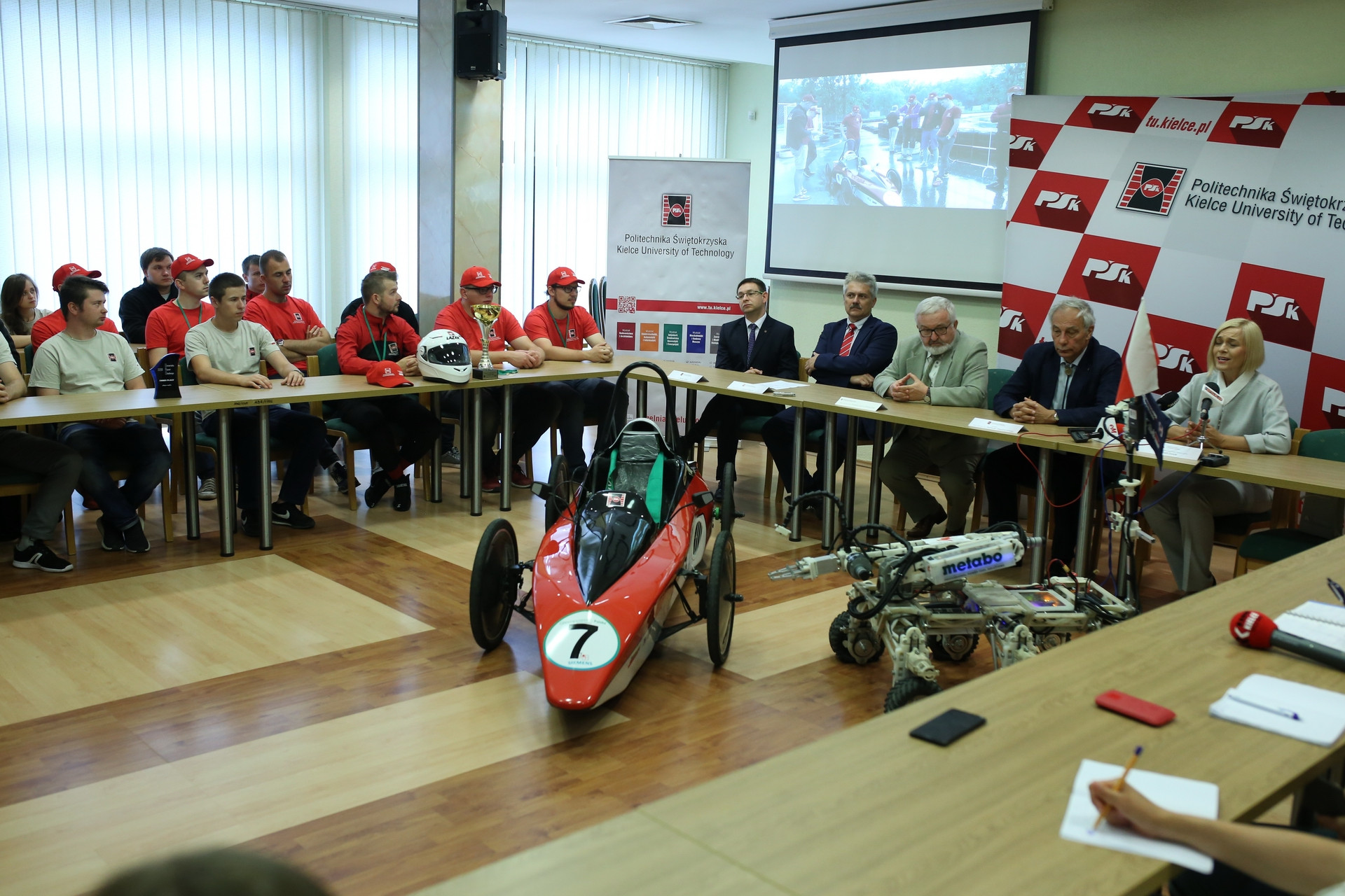 Spotkanie z udziałem studentów i wykładowców Politechniki Świętokrzyskiej. Na stole wyścigowy bolid i robot.