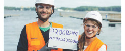 Dwóch pracowników Skanska w kamizelkach odblaskowych trzyma tabliczkę z napisem "PROGRAM AMBASADORÓW" i symbol kciuka w górę.