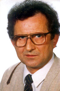 Portret mężczyzny w okularach. Ma na sobie jasną marynarkę i krawat.