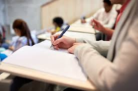Osoba pisze w notatniku podczas wykładu. W tle inni studenci siedzą w amfiteatrze.