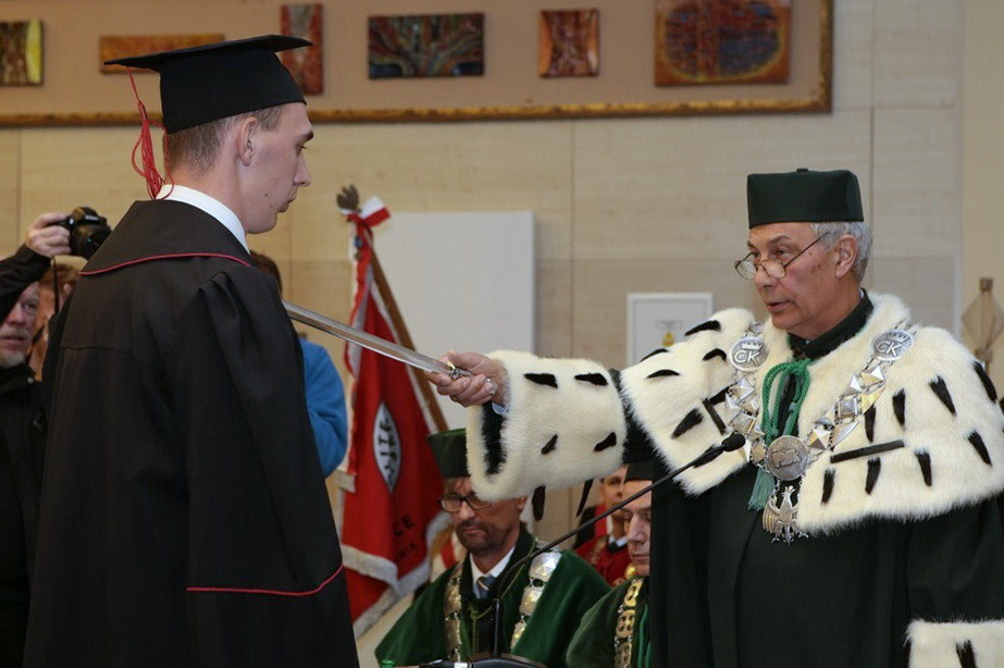 Uroczystość wręczenia nagród. Mężczyzna w todze i birecie otrzymuje miecz od starszego mężczyzny w ceremonialnym stroju.