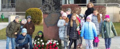 Dzieci i opiekunka przy pomniku. W grupie są chłopcy i dziewczynki w zimowych ubraniach.