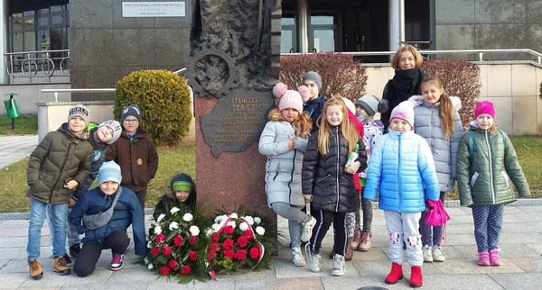 Dzieci i opiekunka przy pomniku. W grupie są chłopcy i dziewczynki w zimowych ubraniach.