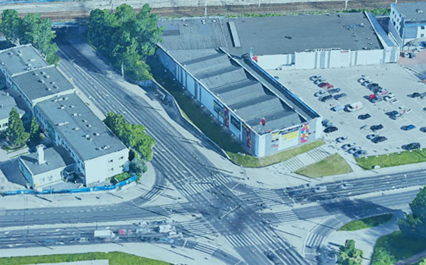 Widok z lotu ptaka na skrzyżowanie dróg z budynkami i parkingiem. Kluczowe obiekty: budynki, samochody, droga.