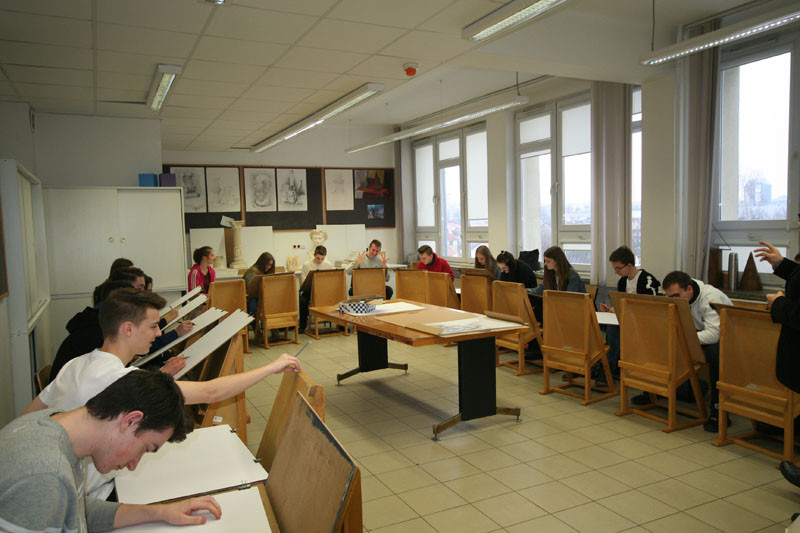 Uczniowie rysują przy sztalugach w pracowni plastycznej. W tle wiszą rysunki i rzeźby.