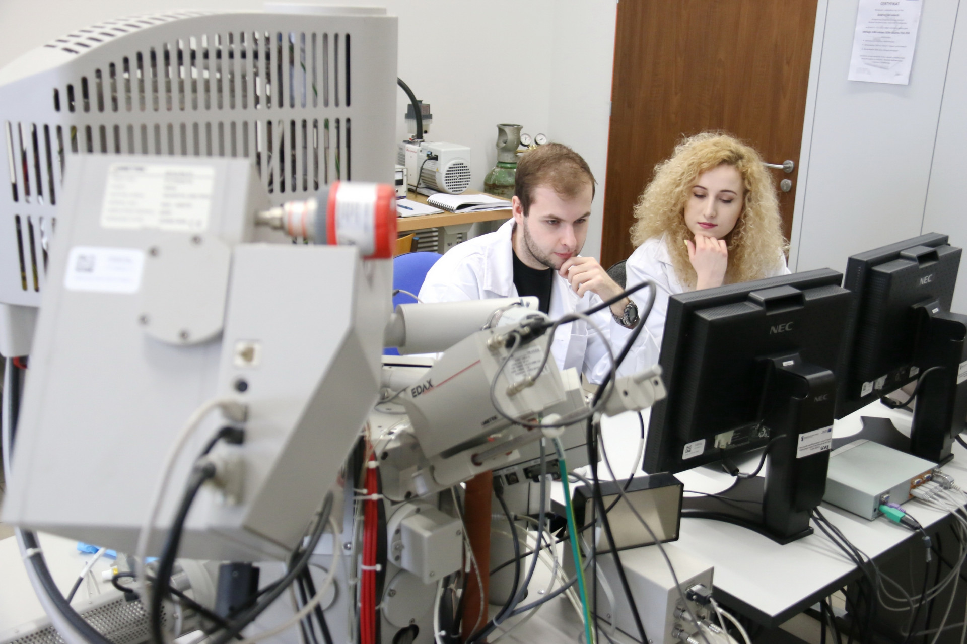 Dwoje naukowców w białych fartuchach pracuje przy skomplikowanej aparaturze laboratoryjnej i monitorach.