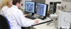 Dwie osoby w białych fartuchach pracują przy mikroskopie elektronowym. Na monitorach widoczne są obrazy mikroskopowe.