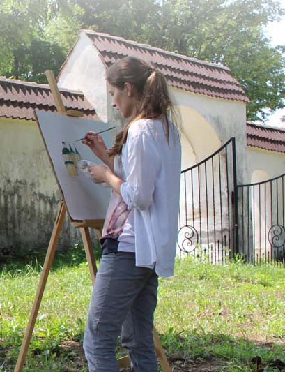 Młoda kobieta maluje na sztaludze na tle zabytkowego muru z bramą.