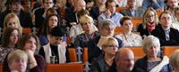 Ludzie siedzą w sali wykładowej, słuchając prelegenta. Widoczne są rzędy krzeseł i twarze publiczności.