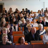Ludzie siedzą w sali wykładowej, słuchając prelegenta. Widoczne są rzędy krzeseł i twarze publiczności.