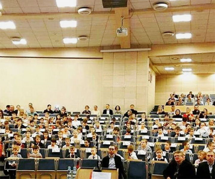 Widownia pełna dzieci i dorosłych siedzących w rzędach. Konferencja lub wydarzenie szkolne.